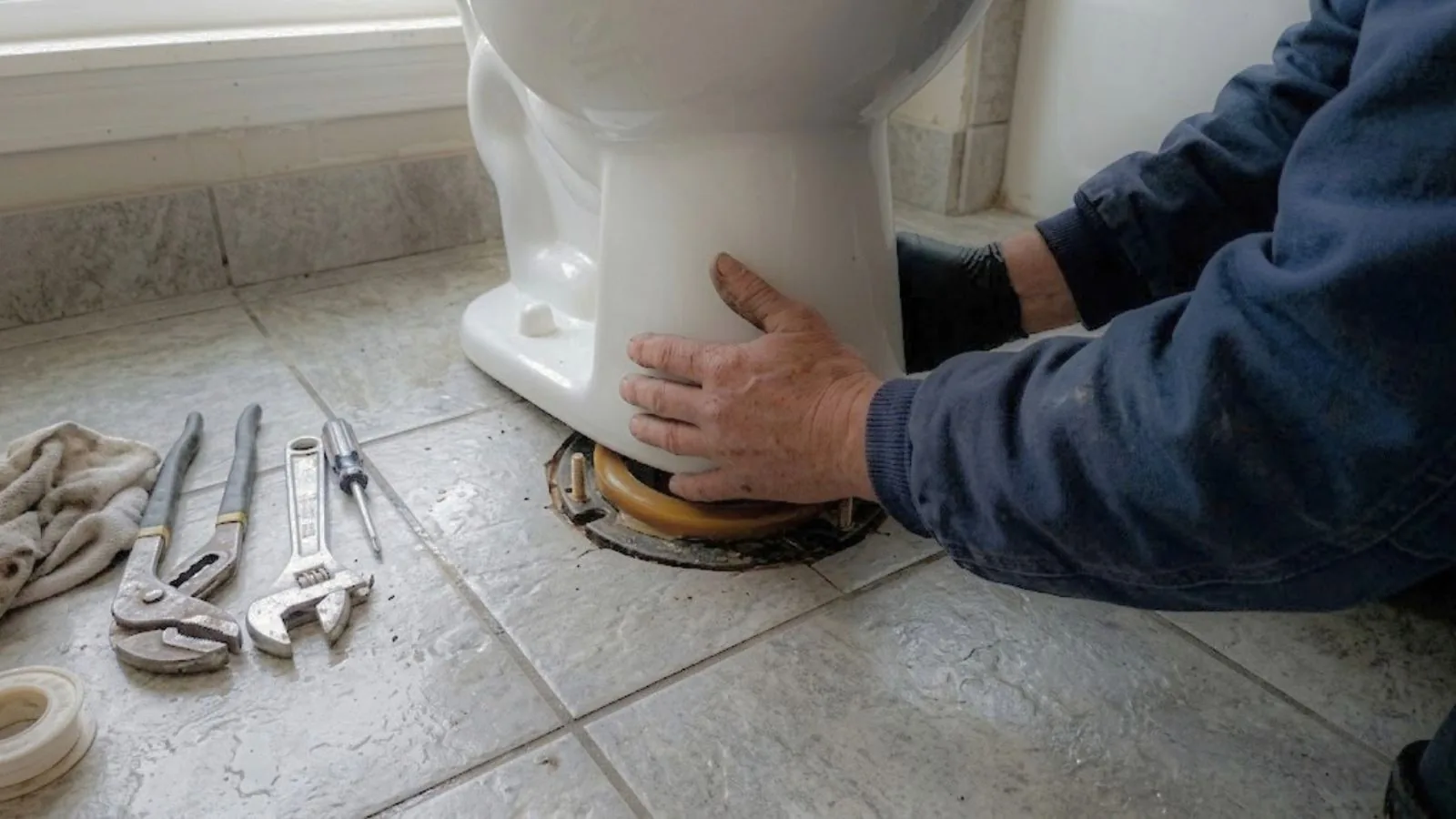 Hands setting a toilet onto a new wax ring with plumbing tools laid out on the floor